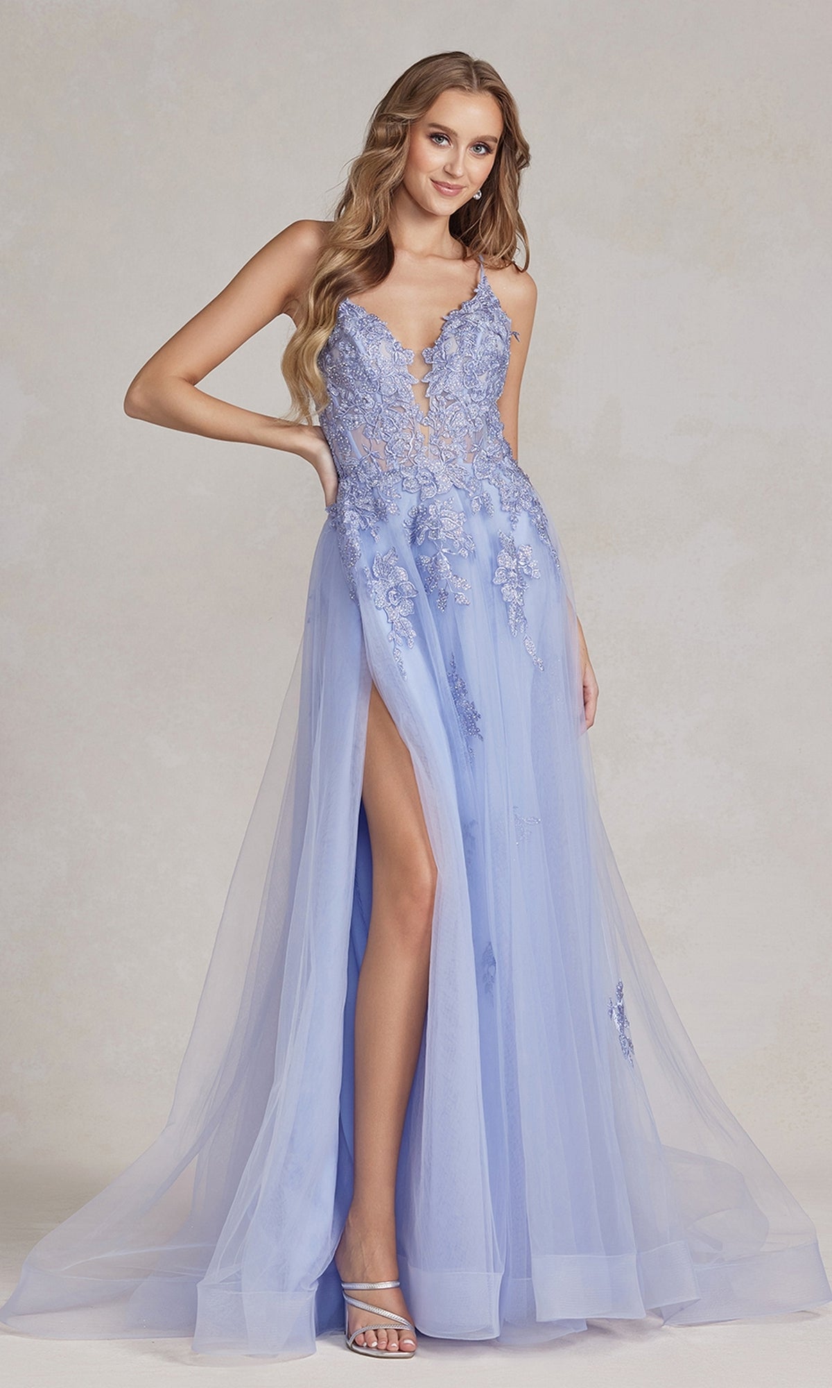A woman poses gracefully against a neutral background in the Open-Back Long Prom Ball Gown with Beads, featuring light blue beaded lace appliqués, spaghetti straps, a sheer overlay, and a thigh-high slit.